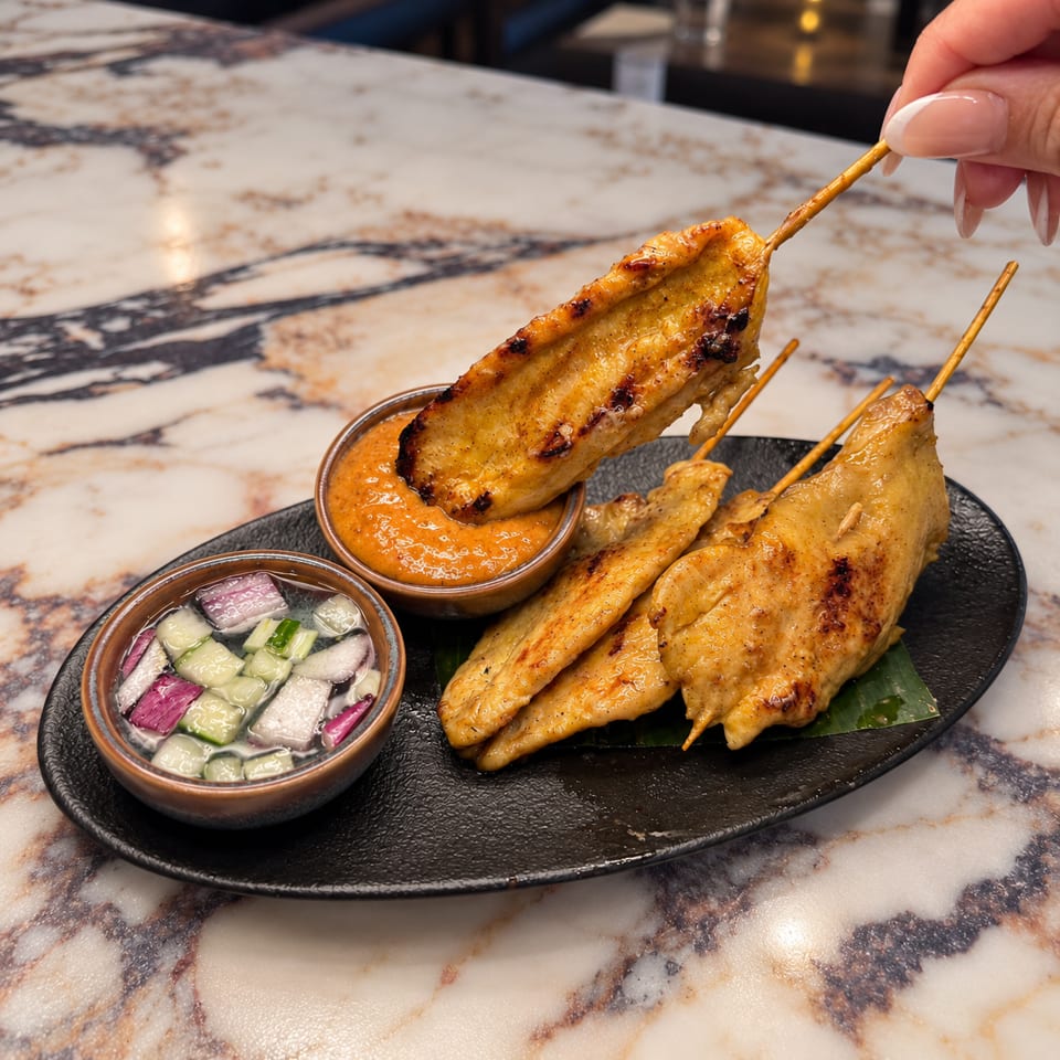 Grilled skewered chicken served with orange dipping sauce and pickled vegetables on a black platter.
