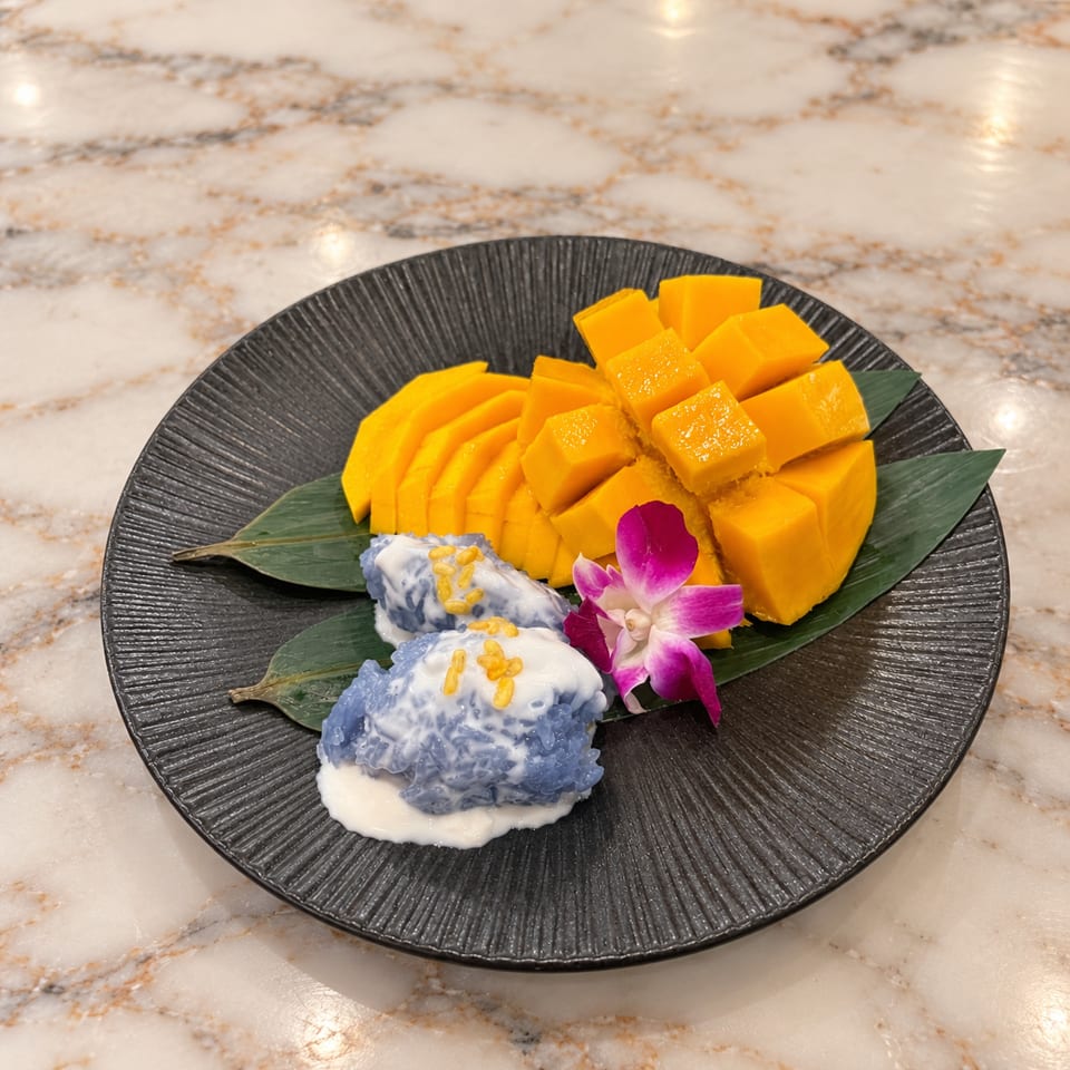 Sliced ripe mango with sticky rice, coconut cream, yellow mung beans, and an orchid on a black plate.