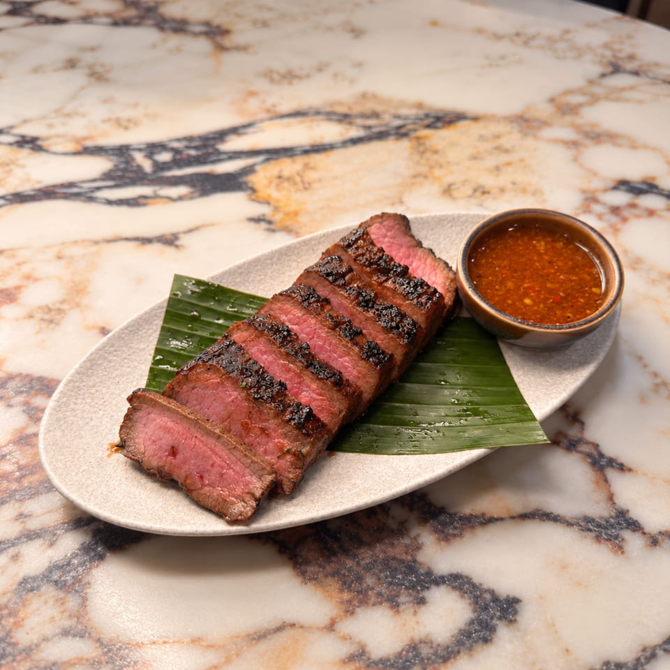 Sliced medium-rare grilled steak rests on a banana leaf with dipping sauce on a ceramic plate.